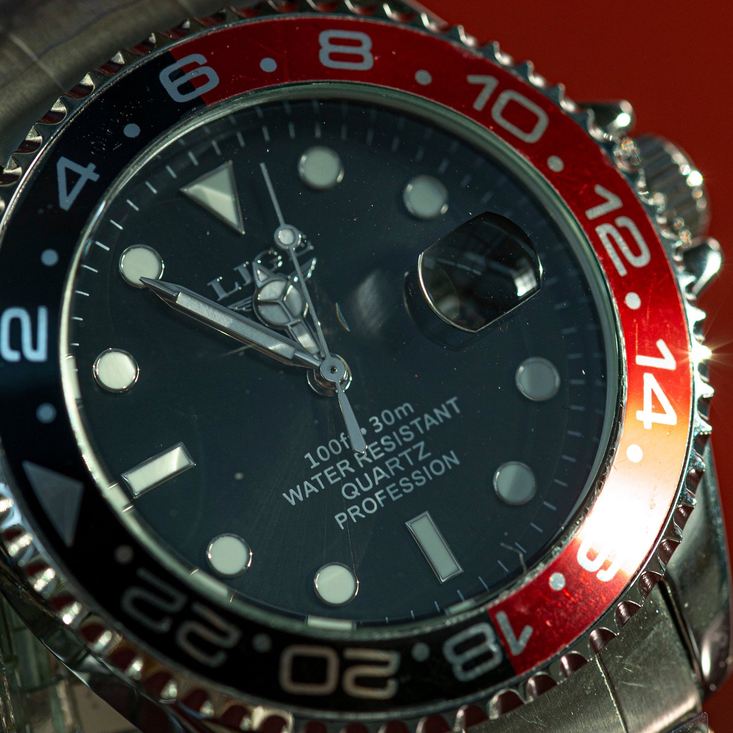 Close-up of a watch with a black face and red and blue bezel on a reflective surface.