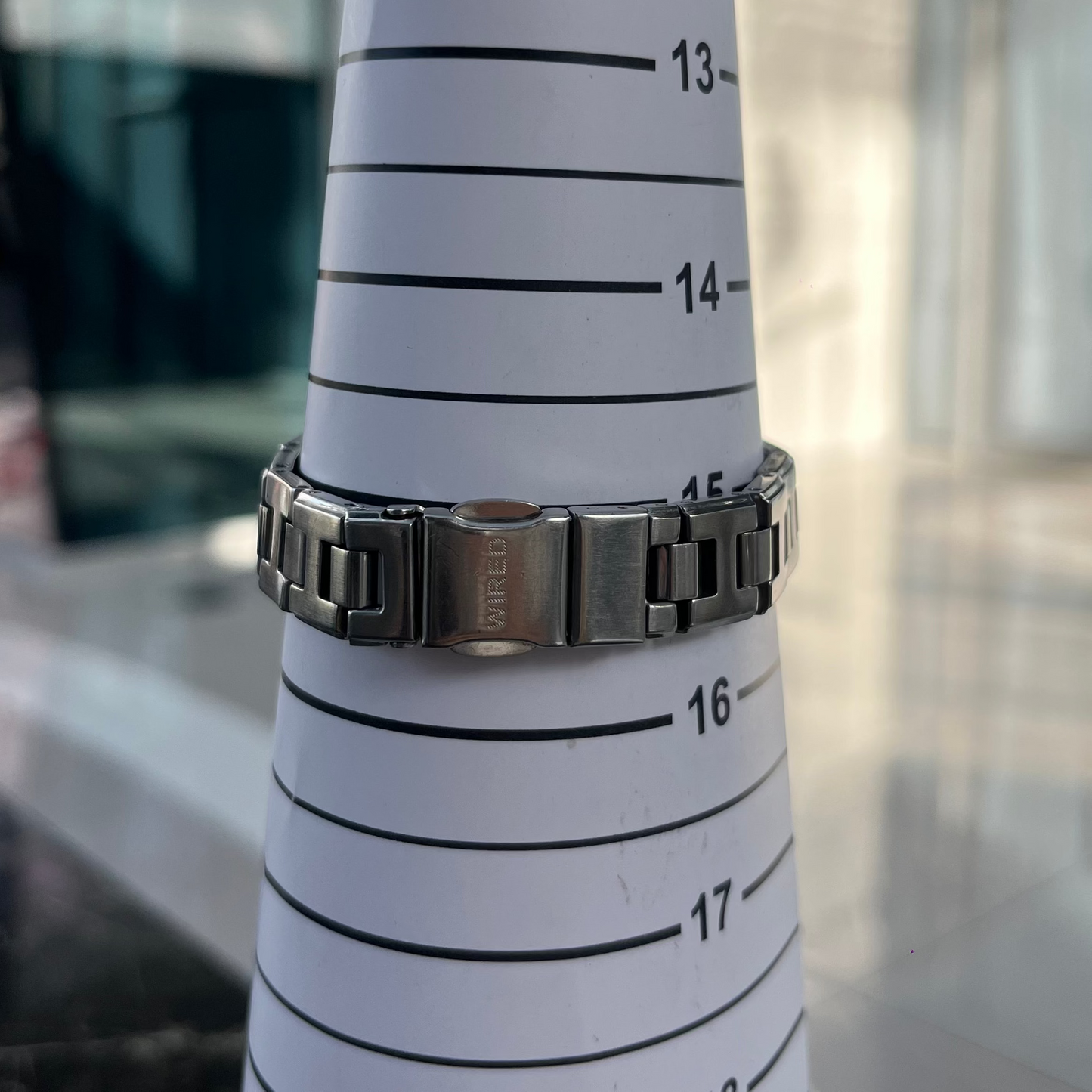 White cylindrical object with black measurement markings and a silver bracelet on top, against a blurred indoor background.