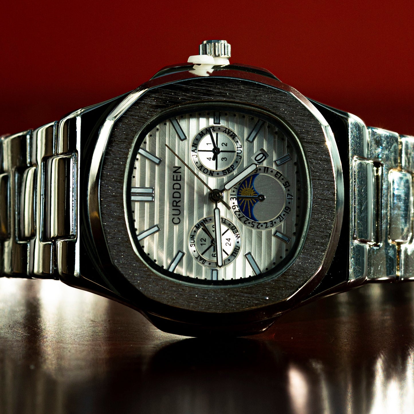 Silver watch with detailed face on a reflective surface against a red background